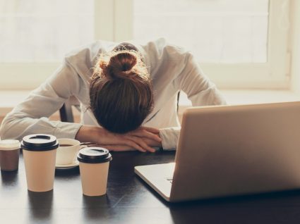 mulher com cabeça na secretária e vários copos de café espalhados pela mesa