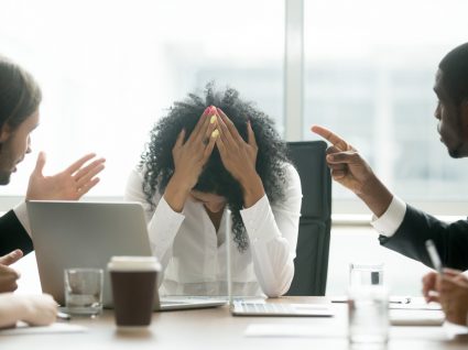 mulher com mãos na cabeça de desepero com dois colegas a falar com ela e apontar dedo