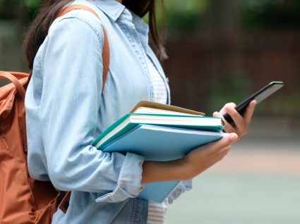 aluna com mochila às costas segura livros numa mão e telefone na outra