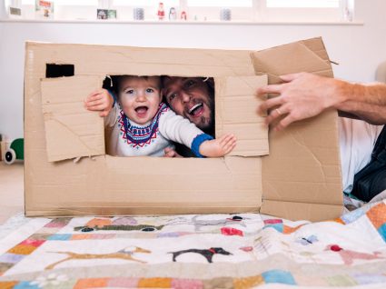 pai e filho bebé a brincar numa casa feita de papel cartão